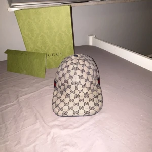 Beige blå keps från Gucci - Snygg beige keps från Gucci med det klassiska GG-mönstret. Kepsen har en justerbar rem baktill och detaljer i rött och blått på sidan. Perfekt för att ge din outfit en lyxig touch. Nypris 5650 kr. PRIS KAN ÄVEN DISKUTERAS (kepsen är Väldigt bra skick)
