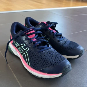 Blå och rosa löparskor från Asics - Säljer ett par snygga Asics GT-1000 löparskor i blått med rosa och mintgröna detaljer. Skorna har snörning och en bekväm passform, perfekta för löpning och träning. De är tillverkade i syntetmaterial med en stabil sula för bra grepp. Storleken är 39 men känns som 38.
