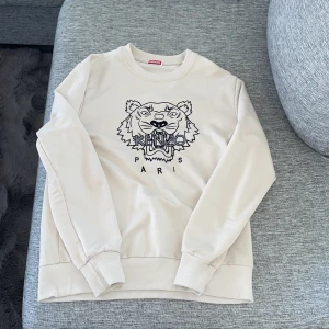 Beige sweatshirt från Kenzo - Säljer en beige sweatshirt från Kenzo med ett stort tigertryck och texten 'Paris' på framsidan. Tröjan har långa ärmar och ribbade muddar vid ärmslut och nederkant. Perfekt för en stilren och bekväm look.