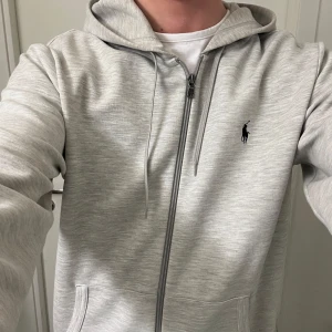Grå hoodie från Ralph Lauren - Säljer en stilren grå hoodie från Ralph Lauren med dragkedja och det klassiska logotypen på bröstet. Perfekt för en avslappnad look. Storlek M, 188cm!