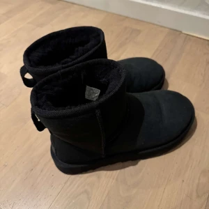 Uggs - Säljer ett par svarta uggs med mjukt foder. De är perfekta för kalla dagar och har en enkel design med rund tå. Skorna är tillverkade i ett material som liknar mocka och har en dragflik baktill för enkel påtagning. 