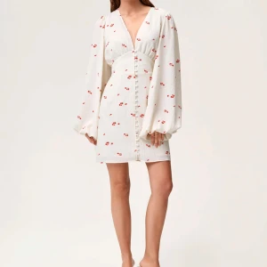 Vit kortklänning med blommönster - Endast använd 2 gånger. Säljer en söt vit kortklänning med små röda blommor. Klänningen har långa puffärmar och en v-ringad design med knappar framtill. Perfekt för sommarens alla tillfällen!