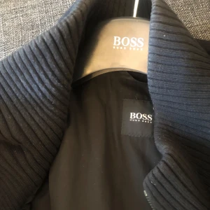 Svart rock från Hugo Boss - Elegant svart rock från Hugo Boss med ribbad krage och knappar framtill. Rocken har flera praktiska fickor med dragkedja på insidan, vilket ger både stil och funktionalitet. Storlek 056 som passar lite större män. Pris kan diskuteras vid en snabb affär 