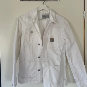 Vit overshirt från Carhartt - Säljer en stilren vit overshirt från Carhartt med knappar framtill och två stora fickor. Jackan har en klassisk krage och en liten logotyp på bröstfickan. Perfekt för lager-på-lager-stil.
