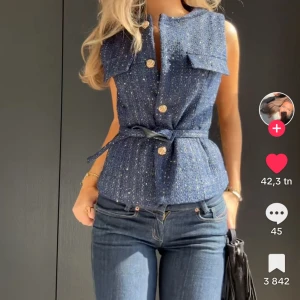 Blå glittrig väst med bälte - Helt ny snygg blå glittrig väst med knappar och bälte i midjan. Perfekt för att ge en extra touch till din outfit. Västen har en elegant design med en figursydd passform.