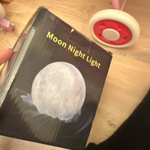Moon Night Light led lampa  - Säljer en Moon Night Light som avger ett mjukt sken och har en design som liknar månen. Den kommer med en fjärrkontroll för att ändra färger och en USB-kabel för laddning. Perfekt för att skapa en mysig atmosfär i rummet. Inkluderar även en träställning för att placera lampan på. 10 cm 