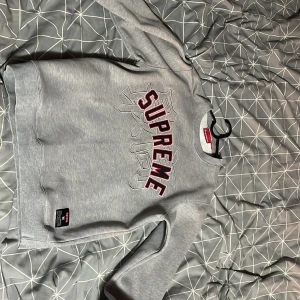 Grå sweatshirt från Supreme - Säljer en grå sweatshirt från Supreme med stort tryck i rött och svart på framsidan. Tröjan har långa ärmar och en klassisk rund halsringning. Perfekt för en avslappnad stil.