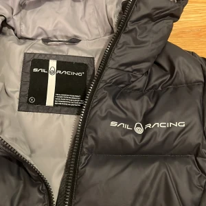 Svart dunjacka från Sail Racing - Fantastiskt skön NY svart dunjacka från Sail Racing i storlek S. Jackan har logoan på bröstet och baksidan av huvan. Perfekt för kyliga dagar och att den går ner längre i bak. Här gärna  av er om frågor eller funderingar!😇❄️NYPRIS  3500kr