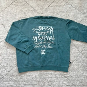 Grön sweatshirt från Stüssy - Säljer en grön sweatshirt från Stüssy med tryck på både fram- och baksidan. Tröjan har långa ärmar och en rund halsringning. Perfekt för en avslappnad stil.