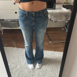 Blå jeans från Anya Jeans - Säljer ett par blå jeans från Anya Jeans med snygga broderade detaljer på bakfickorna. De är låg midjade bootcut och har två hål på framsidan.Köpta i usa så storleken är oklar men jag skulle säga att de passar M. Midjemått:39cm