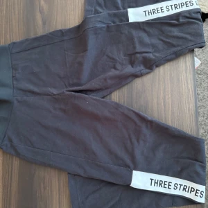 Svarta leggings från Adidas - Snygga svarta leggings från Adidas med texten 'THREE STRIPES' längs benen. Perfekta för träning eller en avslappnad dag. Tillverkade i ett mjukt och stretchigt material för bästa komfort.