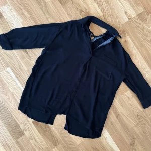 Svart blus med knytband - Snygg svart blus med knytband i halsen och en bröstficka. Blusen har trekvartsärmar och en avslappnad passform. Perfekt för en stilren look.