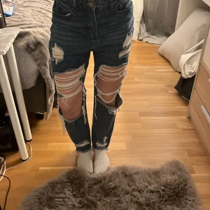 Blå slitna jeans - Säljer ett par blå jeans med rejält slitna detaljer och hål på både fram- och baksida. De har en hög midja och en straight passform. Perfekta för en avslappnad och trendig look.