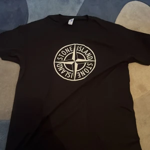 Svart t-shirt från Stone Island - Snygg svart t-shirt från Stone Island med deras ikoniska logga tryckt på framsidan. Perfekt för en stilren och avslappnad look.