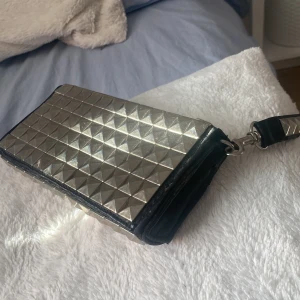 Silvrig plånbok från Gina Tricot - Snygg och stilren plånbok från Gina Tricot i silver med pyramidformade nitar. Den har en avtagbar handledsrem och stängs med dragkedja. Perfekt för att ge en edgy touch till din outfit.