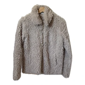 Beige teddyjacka från Lindex - Mysig beige teddyjacka från Lindex för barn men fungerar bra som S i damstorlek. Jackan har en dragkedja framtill och är perfekt för kyligare dagar. Den fluffiga texturen ger en trendig look och håller dig varm och bekväm.