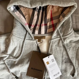 Grå hoodie från Burberry - Säljer en stilren grå hoodie från Burberry med klassiskt rutigt mönster i luvan. Tröjan har en dragkedja framtill och ett broderat emblem på bröstet. Perfekt för en avslappnad och trendig look.