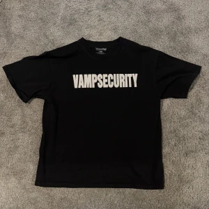 Svart t-shirt med tryck 'Vampsecurity' - Säljer en svart t-shirt från NDCRP med det coola trycket 'Vampsecurity' i vitt på framsidan. Tillverkad i 100% bomull för en skön känsla. Perfekt för dig som vill ha en unik stil. 🖤