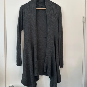 Grå ribbstickad kofta från KappAhl - Säljer en stilren grå kofta från KappAhl. Den är ribbad och har en öppen design med långa ärmar. Perfekt för lager-på-lager under kyligare dagar. Materialet är en blandning av bomull och nylon vilket ger en mjuk och bekväm känsla.