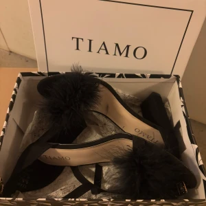 Svarta klackskor med pälsdetalj från Tiamo - Eleganta svarta klackskor från Tiamo med en unik pälsdetalj på ovansidan. Skorna har en öppen tå och en bekväm klack, perfekt för att ge en stilfull touch till din outfit.