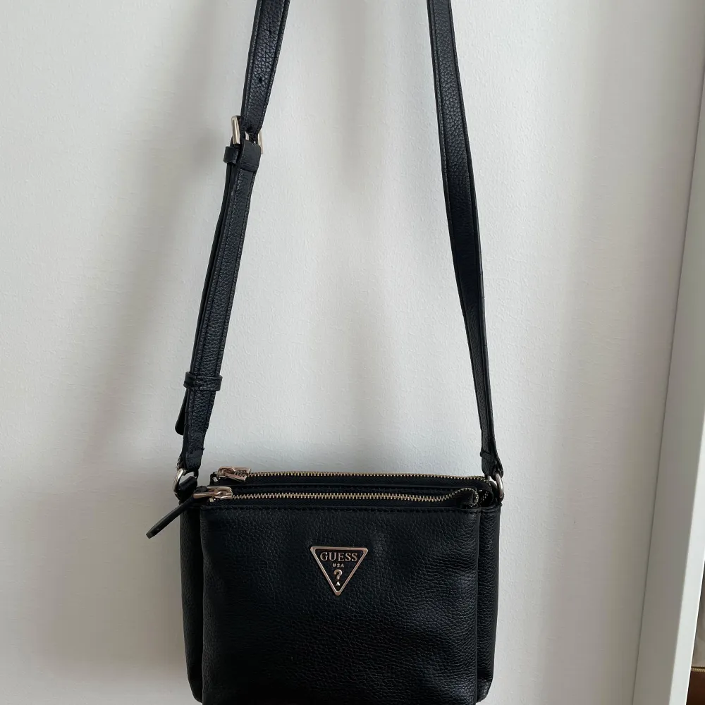 Snygg svart axelväska från Guess med justerbar rem och dragkedja. Väskan har en stilren design med Guess-logotypen i metall på framsidan. Perfekt för att bära dina nödvändigheter med stil. Liten skada i nedre hörnet och 1 blixtlåshuvud saknas. Laukut & Käsilaukut.
