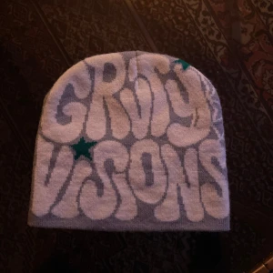 Grå mössa med text och stjärnor - Snygg grå mössa med texten 'GROOVY VISIONS' i stora vita bokstäver och gröna stjärnor. På baksidan finns ett coolt spindelnätsmönster. Perfekt för att hålla stilen i kylan!