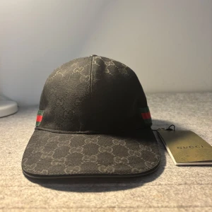 Svart gucci keps - Snygg svart keps från Gucci med diskret mönster och klassiska ränder i rött och grönt på sidan. Perfekt för att ge din outfit en lyxig touch. Storlek ”S”