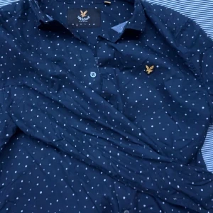 Mörkblå prickig skjorta från Lyle & Scott - Säljer en stilren mörkblå skjorta från Lyle & Scott med vita prickar och en broderad logga på bröstet. Skjortan har långa ärmar och knappar framtill. Perfekt för en snygg och avslappnad look.