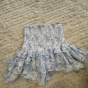 Blåvit kjol från Gina Tricot - Säljer en söt blåvit kjol från Gina Tricot med volangdetaljer. Kjolen har ett blommigt mönster och är perfekt för sommardagar. Den har en elastisk midja för en bekväm passform.