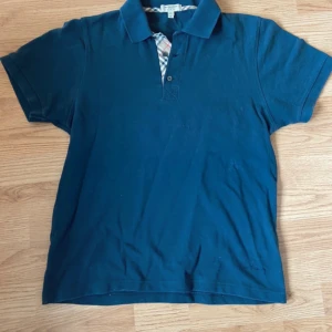 Blå pikétröja från Burberry - Säljer en stilren blå pikétröja från Burberry med klassisk krage och knappslå. Tröjan har ett diskret rutigt mönster på insidan av kragen och knappslån. Perfekt för en avslappnad men ändå elegant look.