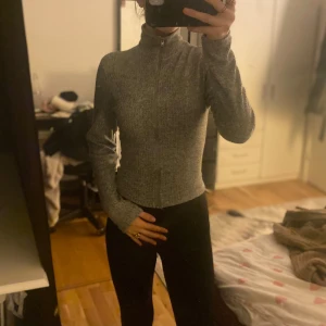 Grå ribbad tröja med dragkedja - Säljer en stilren grå ribbad tröja med dragkedja och hög krage. Perfekt för en casual look. Tröjan har långa ärmar och en tight passform.