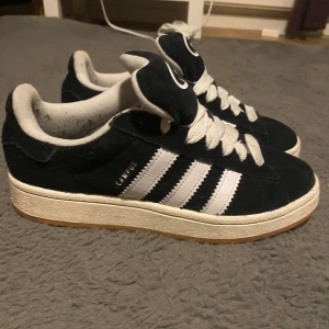Svarta Adidas Campus sneakers - Snygga svarta Adidas Campus sneakers med vita ränder och snörning. Skorna har en klassisk design med gummisula och är perfekta för en avslappnad stil.