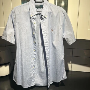 Ljusblå kortärmad skjorta från Ralph Lauren - Säljer en stilren ljusblå kortärmad skjorta från Ralph Lauren med klassisk krage och knappar framtill. Skjortan har en broderad logotyp på bröstet som ger en elegant touch. Perfekt för en avslappnad men ändå uppklädd look.