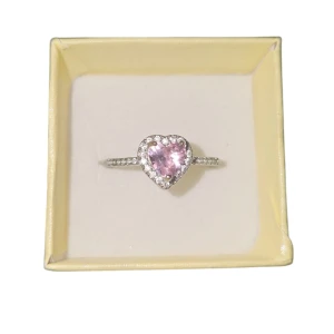 Hjärtaformad ring med rosa sten - Elegant ring med en hjärtaformad rosa sten i mitten, omgiven av glittrande stenar. Perfekt för att ge en romantisk touch till din stil. US 7.5 eller 17.7 mm, okänt material 