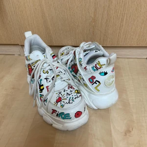  Buffalo - Unika vita sneakers från Buffalo med färgglatt graffiti-mönster. Skorna har en chunky sula och snörning framtill. Perfekta för att ge din outfit en cool och lekfull touch.