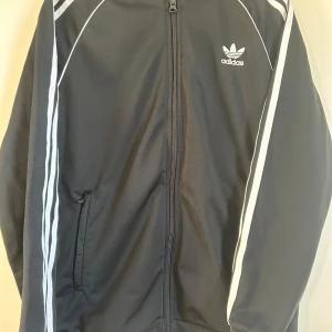 Svart träningsjacka från Adidas 176cl/XL  - Snygg svart träningsjacka från Adidas med klassiska vita ränder längs ärmarna och dragkedja framtill. Två sidfickor med dragkedja. Jackan har en normal passform och är perfekt för sportiga aktiviteter. Adidas-loggan är broderad på bröstet. Nästan oanvänd så i mkt bra skick!