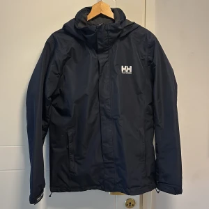 Svart vindjacka från Helly Hansen - Säljer en svart vindjacka från Helly Hansen med huva och dragkedja. Jackan har en stilren design med Helly Hansen-logga på bröstet och nacken. Perfekt för blåsiga dagar.