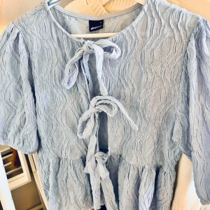 Ljusblå kofta från Gina Tricot - Charmig ljusblå kofta med knytning framtill. Den har en krinklad textur och korta ärmar. Perfekt för en somrig look!