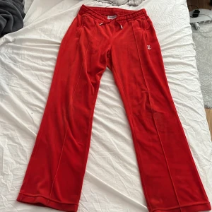 Röda mjukisbyxor från Juicy Couture - Snygga röda mjukisbyxor från Juicy Couture med glittrande text på baksidan. Byxorna har en elastisk midja med snörning och är tillverkade i ett mjukt material för extra komfort. Perfekta för en avslappnad stil.