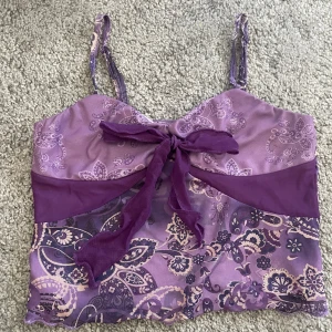 Lila mönstrad linnetopp - Säljer en lila mönstrad linnetopp med justerbara axelband och en söt knytdetalj framtill. Toppen har ett paisleymönster i olika nyanser av lila och beige. 