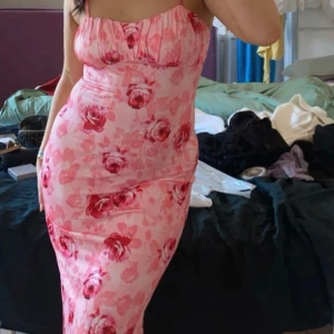 Rosa blommig klänning - Säljer en söt rosa klänning med blommigt mönster. Klänningen har smala axelband och en figurnära passform som framhäver formerna. Perfekt för sommarens festligheter!