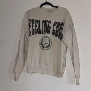 Vit sweatshirt från Bershka - Säljer en vit sweatshirt från Bershka med texten 'FEELING COOL' i svart på framsidan. Tröjan har långa ärmar och en rund halsringning. Perfekt för en avslappnad stil. Lite missfärgningar annars fin