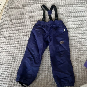 Mörkblå täckbyxor med hängslen från Petite Chérie - Säljer ett par mörkblå täckbyxor från Petite Chérie med justerbara hängslen. Byxorna har elastisk midja och reflexdetaljer i form av hjärtan på benen. Perfekta för kalla dagar.