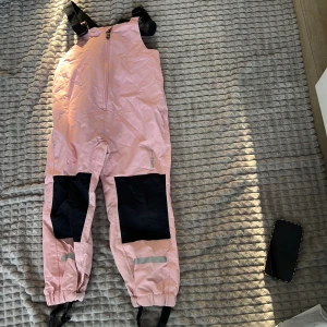 Rosa täckbyxor med svarta detaljer - Säljer ett par rosa täckbyxor med svarta förstärkningar på knäna och justerbara axelband. Byxorna har en dragkedja framtill och reflexdetaljer vid benen. Perfekta för vinteraktiviteter!