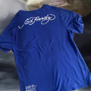 Blå t-shirt från Ed Hardy - Säljer en blå t-shirt från Ed Hardy med ett coolt tryck av en dödskalle och hjärta på framsidan. T-shirten har korta ärmar och är gjord i bomull. Perfekt för dig som gillar en edgy stil!   (Liten fläck på baksidan)