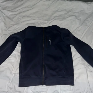 Mörkblå huvtröja från Sail Racing - Säljer en mörkblå huvtröja från Sail Racing med dragkedja framtill. Tröjan har en bröstficka med dragkedja och en bekväm huva. Priset kan diskuteras.