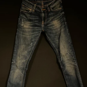 Mörkblå jeans från Nudie Jeans - Snygga mörkblå jeans från Nudie Jeans med slitna detaljer och orange sömmar. Perfekta för en avslappnad stil med en touch av edge. Storlek 26 28