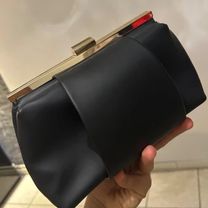 Svart kuvertväska med gulddetaljer - Elegant svart kuvertväska med stilrena gulddetaljer. Väskan har en modern design med en metallisk stängning upptill. Perfekt för att ge en touch av lyx till din outfit.