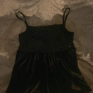 Svart paljettklänning med axelband - Snygg svart blus med paletter på överdelen och smala axelband. Perfekt för en glittrig kväll. Kjoldelen är i ett mjukt material som ger en fin kontrast till den glittriga toppen.