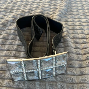 Svart bälte med glittrigt spänne - Snyggt svart bälte med ett stort, glittrigt spänne dekorerat med stora fyrkantiga stenar och mindre gnistrande detaljer. Perfekt för att ge en extra touch till din outfit.